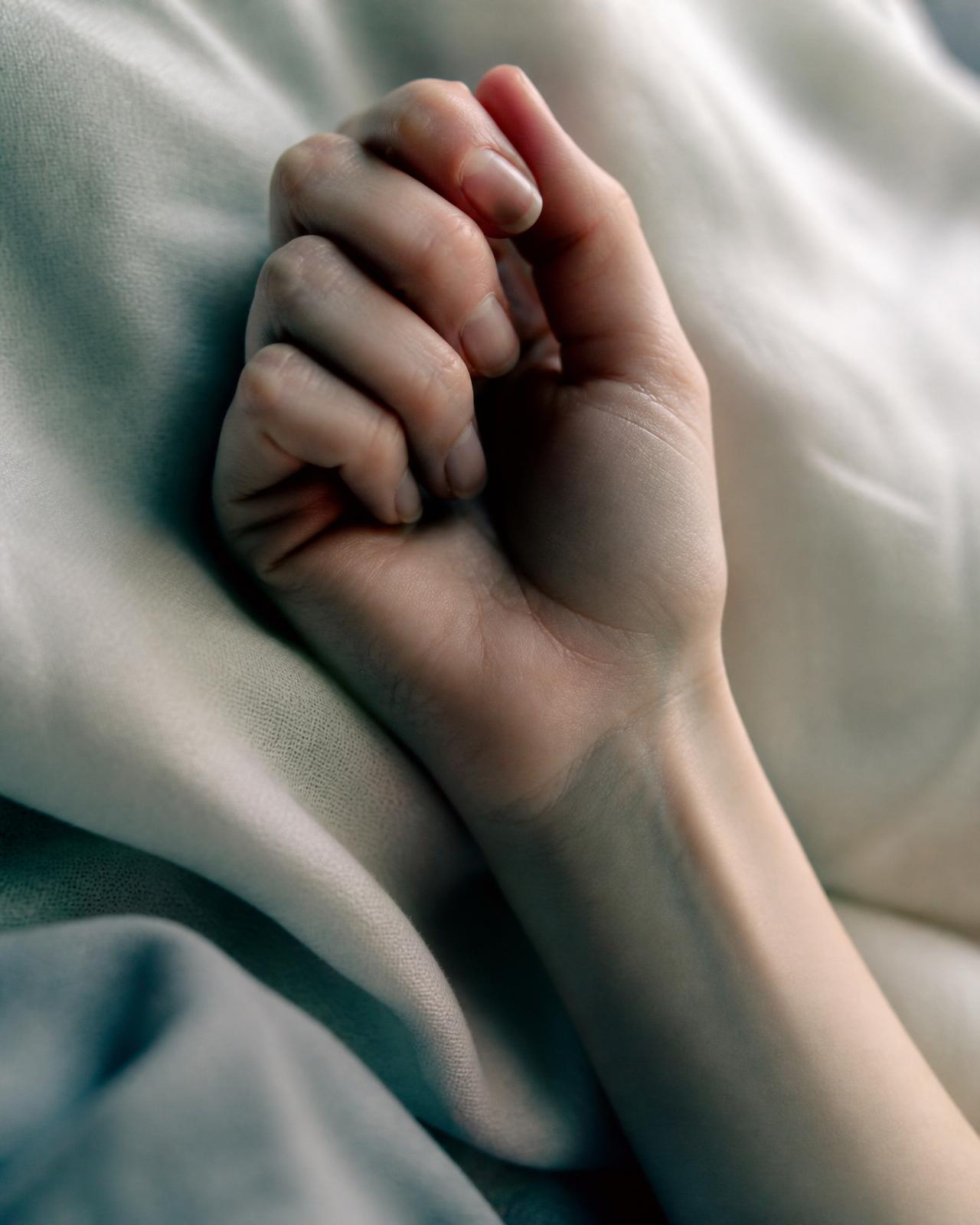 A clenched hand against soft cream linen — the quiet visual of a body in spasm