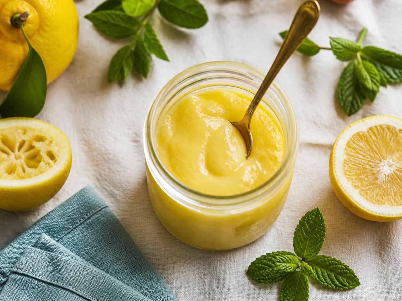 Jar of glossy lemon and mint curd with brass spoon, mint leaves and halved lemons on cream linen
